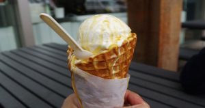 A hand holding a waffle ice-cream cone with vanilla ice-cream and a spoon sticking out of it to the left.