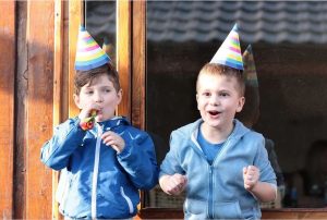 Two little boys standing outside of a building with birthday party hats.