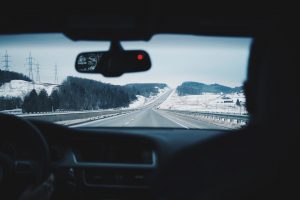 Interior front seat view of the road with snow  and trees on the side from the front seat. 