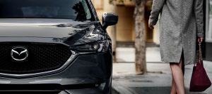 Close view of the front driver side of a gray 2020 Mazda CX-5 parked by a sidewalk with a woman wearing a long gray jacket standing beside it. | Mazda dealer in Ellisville, MO