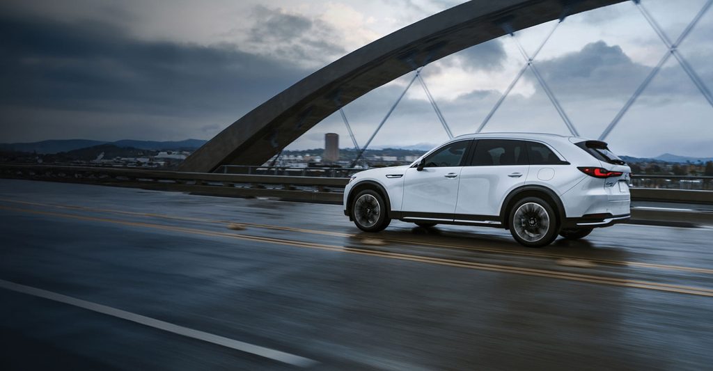 White Mazda CX-90 Performance SUV driving on a wet road with a bridge in the background.
