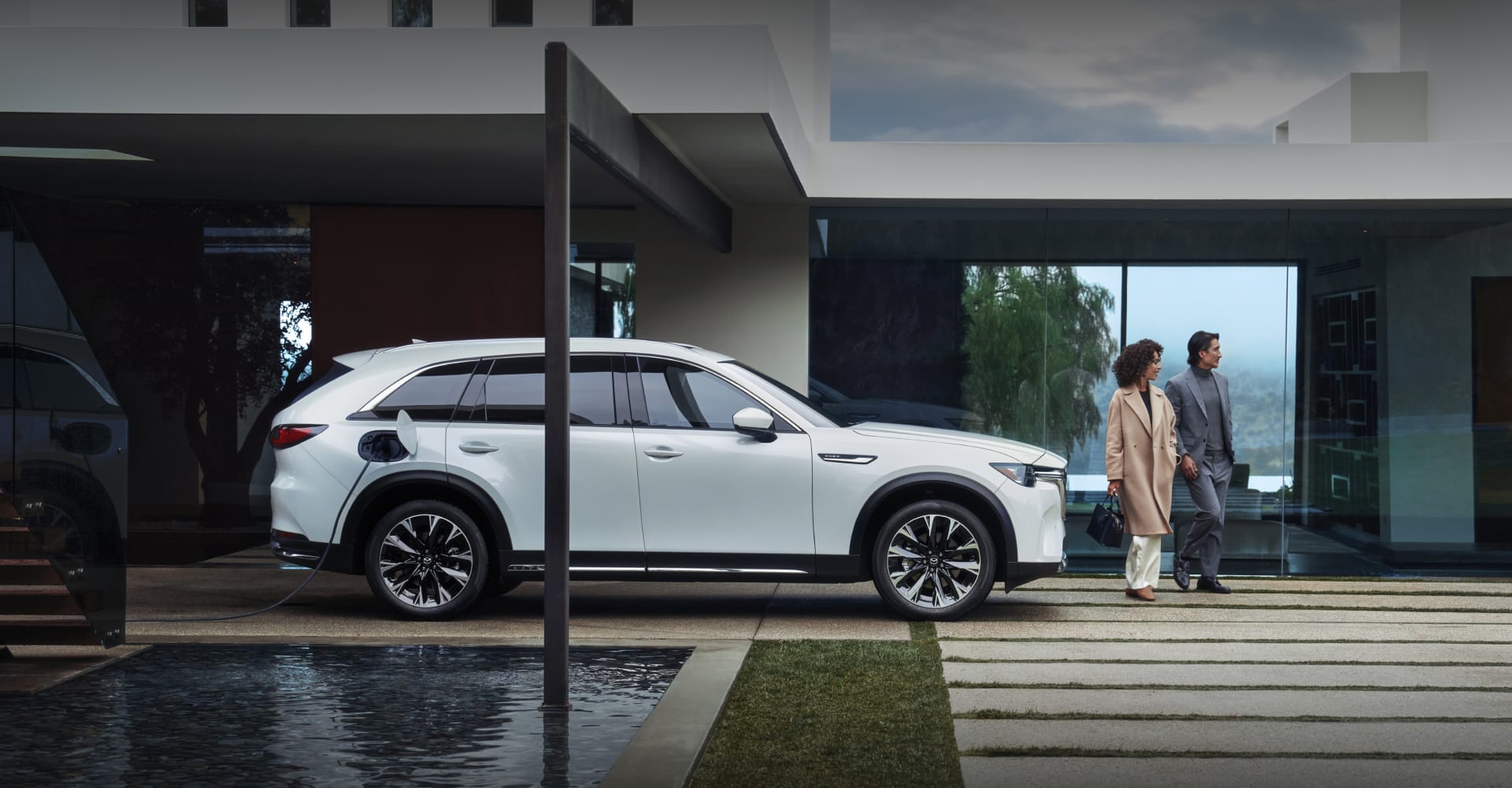 White Mazda CX-90 PHEV parked outside a modern home with people nearby.